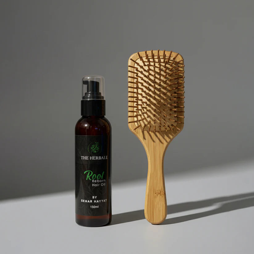 The Herbale Root Reborn Hair Oil 150ml bottle placed next to a wooden paddle hairbrush on a gray background.