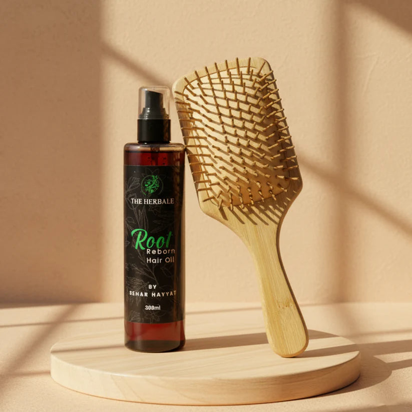 The Herbale Root Reborn Hair Oil 300ml bottle next to a wooden paddle hairbrush on a wooden platform with sunlight streaming in.