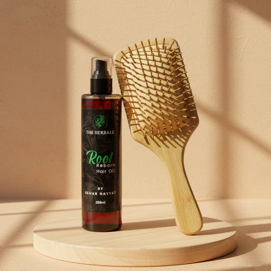 The Herbale Root Reborn Hair Oil 300ml bottle next to a wooden paddle hairbrush on a wooden platform with sunlight streaming in.